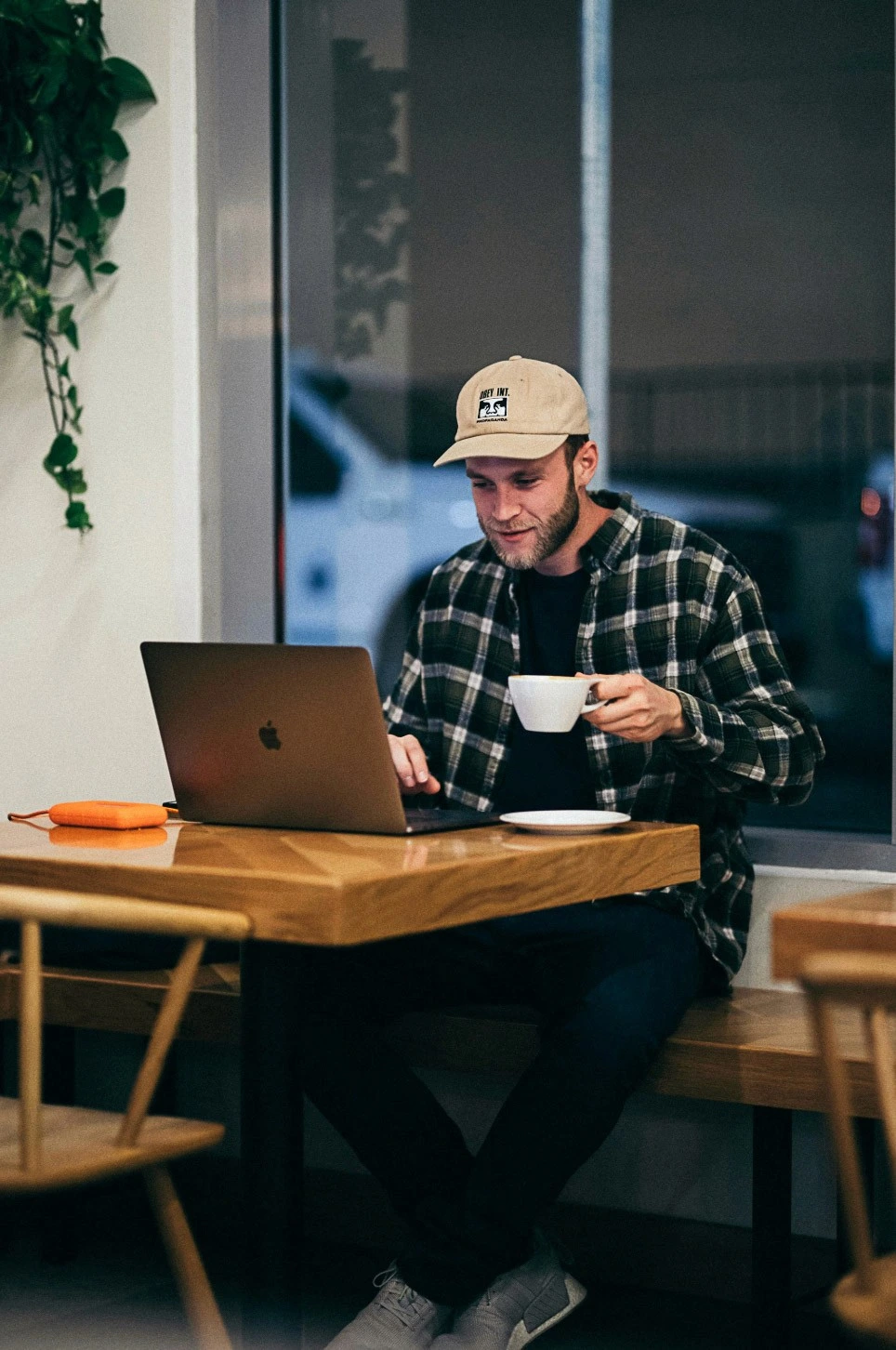 A man wearing a cap and flannel shirt works on his laptop with a coffee in a café – Blursafe can be used anywhere to securely anonymize documents.